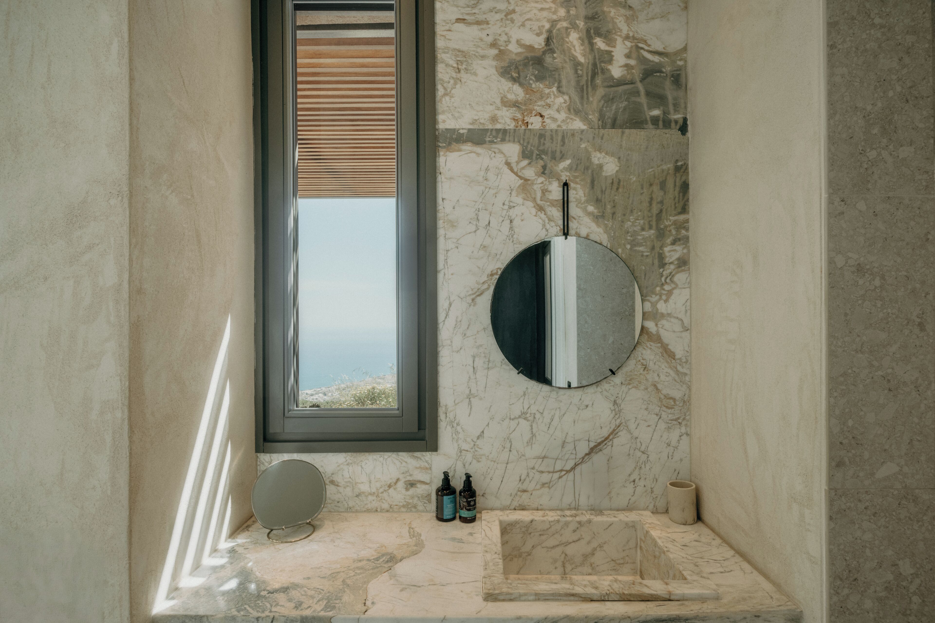 A bathroom with a marble sink, a round mirror, and a window with blinds.