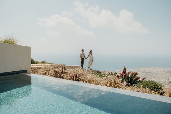 A couple holding hands by a pool with a view of the ocean. - Santorini Sky, Luxury Resort (Santorini)