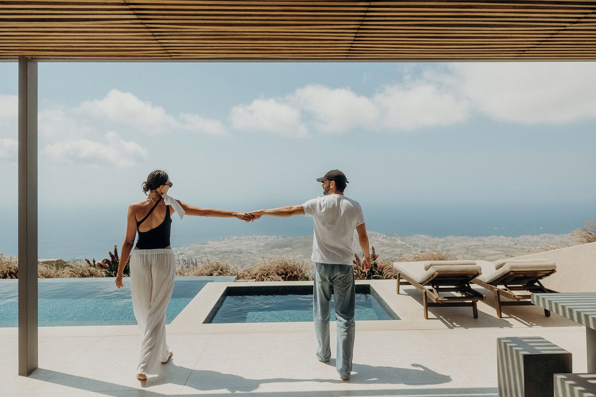 Una pareja tomándose de las manos junto a una piscina con vistas al océano y las colinas.