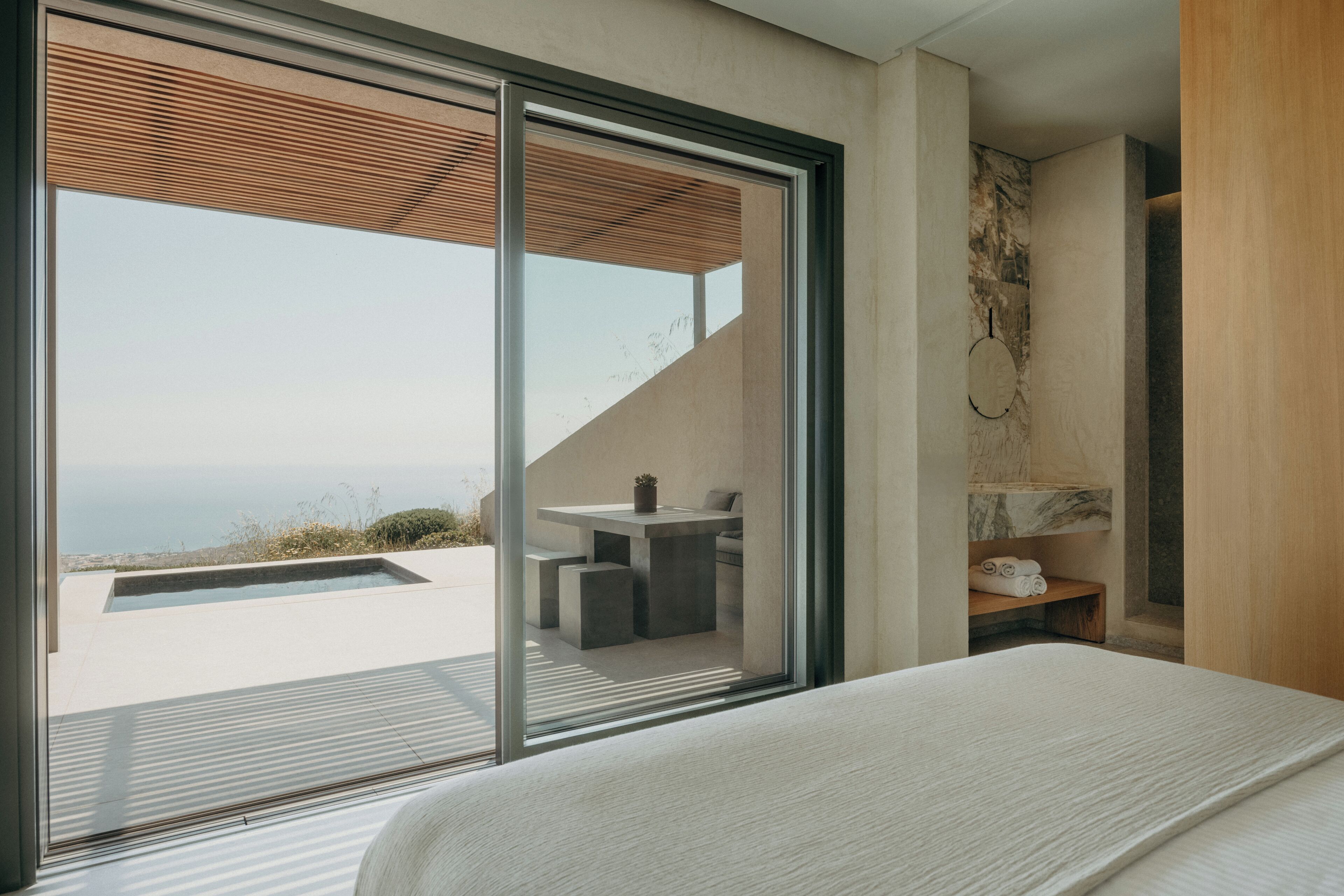 A modern bedroom with a large bed, a sliding glass door leading to a balcony with a pool, and a small seating area with a table and chairs.