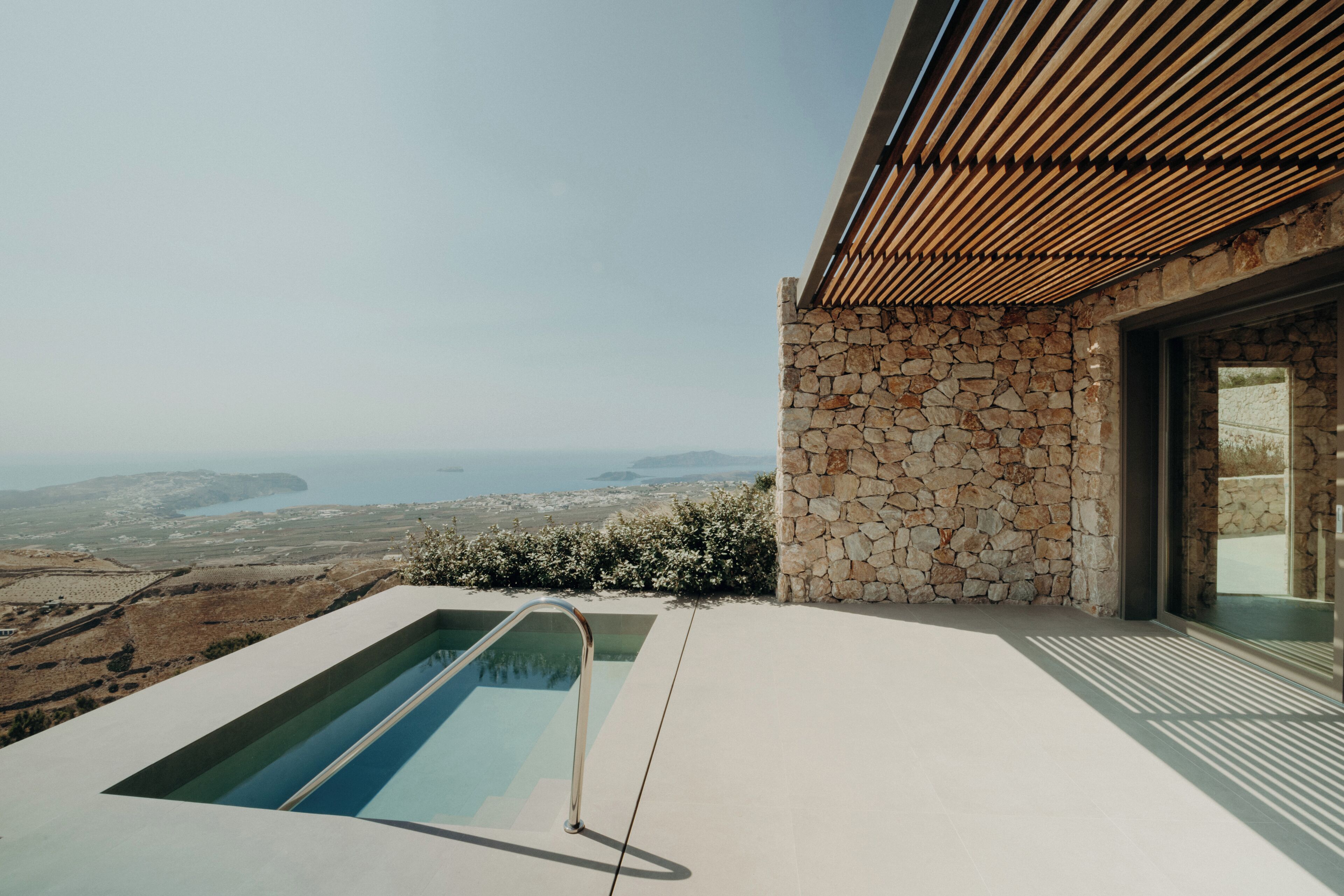 A modern pool with a stone wall and wooden slats on the roof, overlooking a coastal landscape.