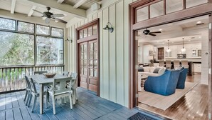 A covered porch with a dining table and chairs, a wooden door, and a view into a modern kitchen with white cabinets and blue chairs. - 2 Minutes to Beach Club. Screened-in Porches, Bikes, Grill. Best Location! (Santa Rosa Beach)
