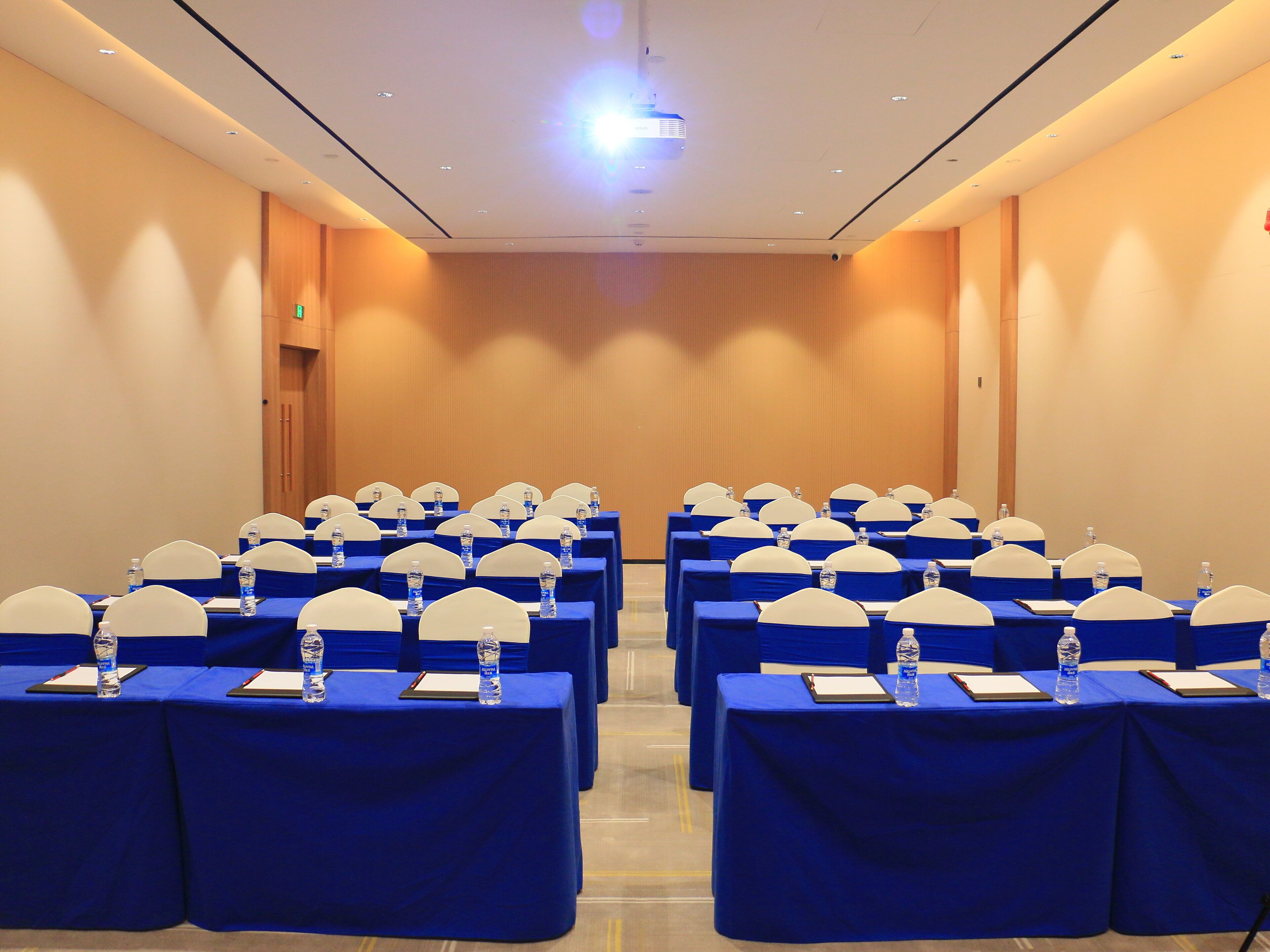 Uma sala de conferências com fileiras de mesas, cada uma com toalha azul, capas brancas para as cadeiras e garrafas de água.