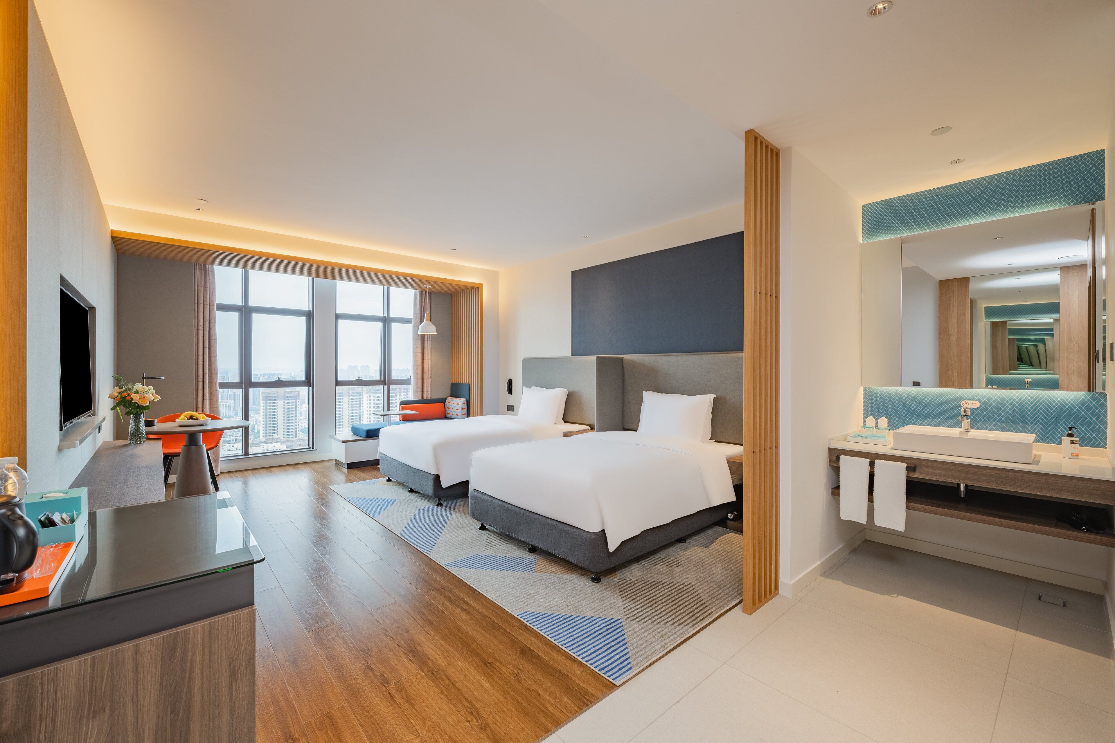 A modern hotel room with a large bed, a desk, a bathroom with a mirror, and a view of the city.