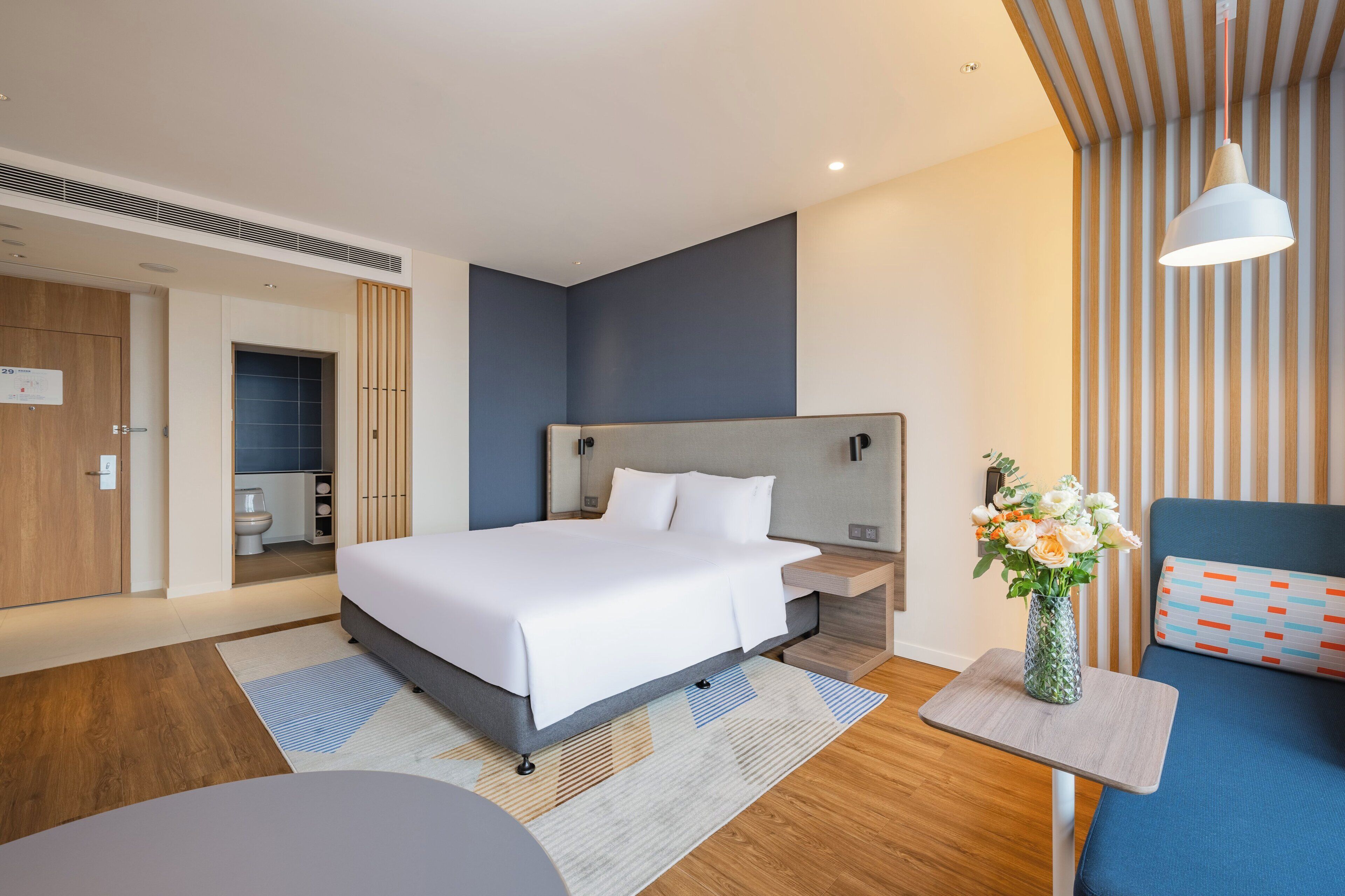 Ein modernes Hotelzimmer mit einem großen Bett, einem Nachttisch, einem kleinen Tisch mit Blumen und einer Sitzecke mit einem blauen Sofa.