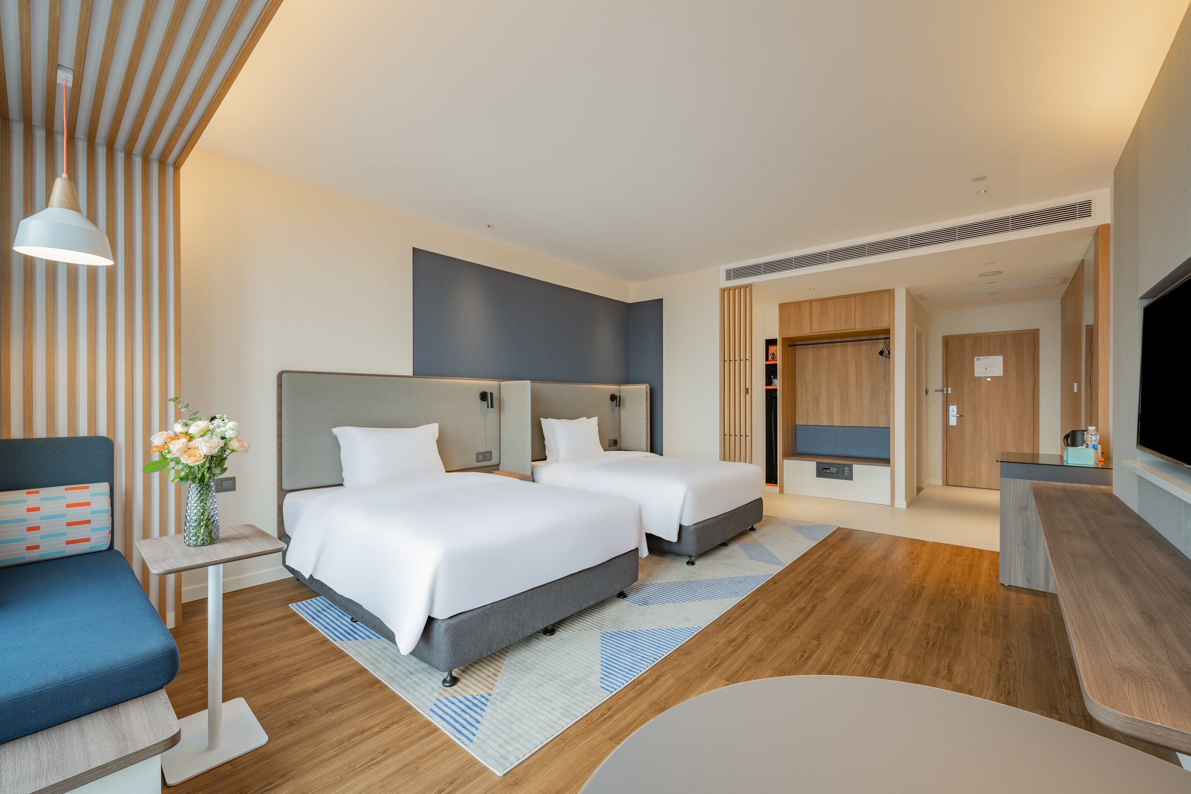 A modern hotel room with two beds, a desk, a TV, and a seating area.