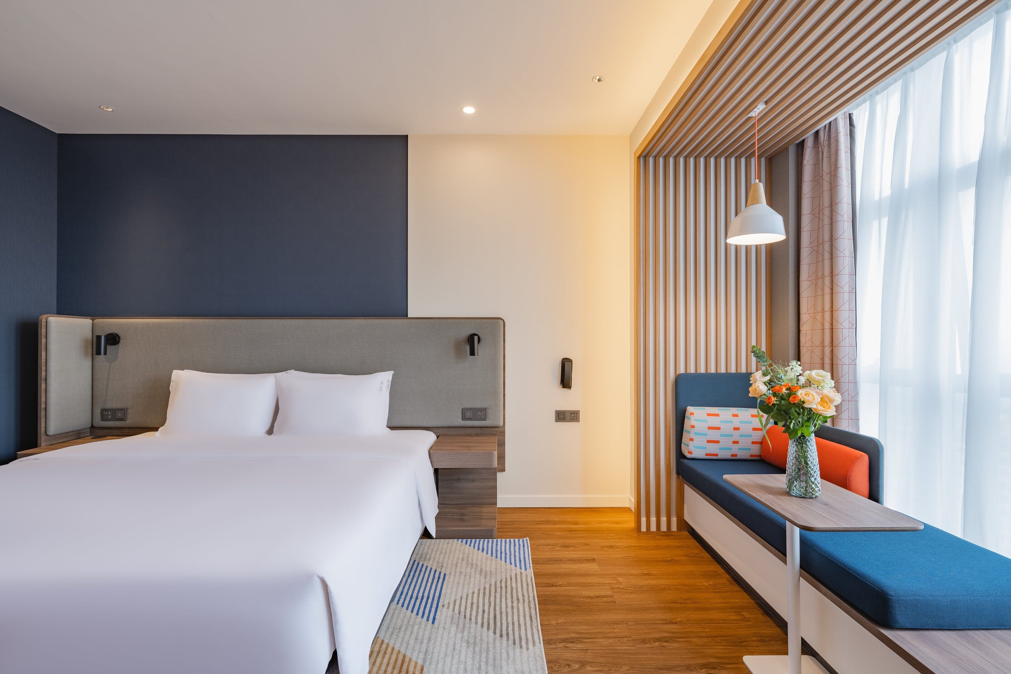 A modern hotel room with a large bed, a seating area with a vase of flowers, and a view through a window with curtains.