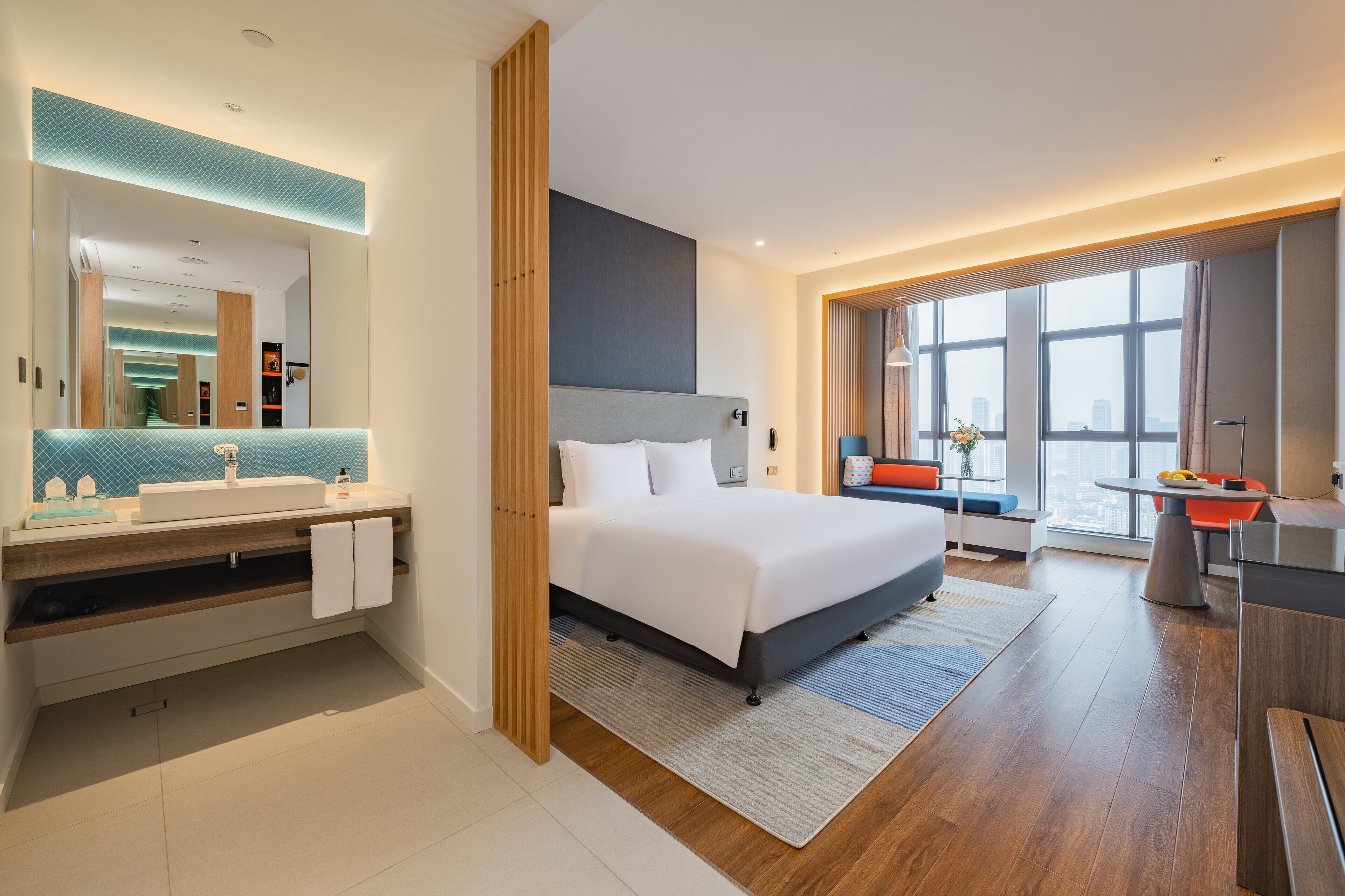 A modern hotel room with a large bed, a bathroom with a vanity, and a view of the city through large windows.