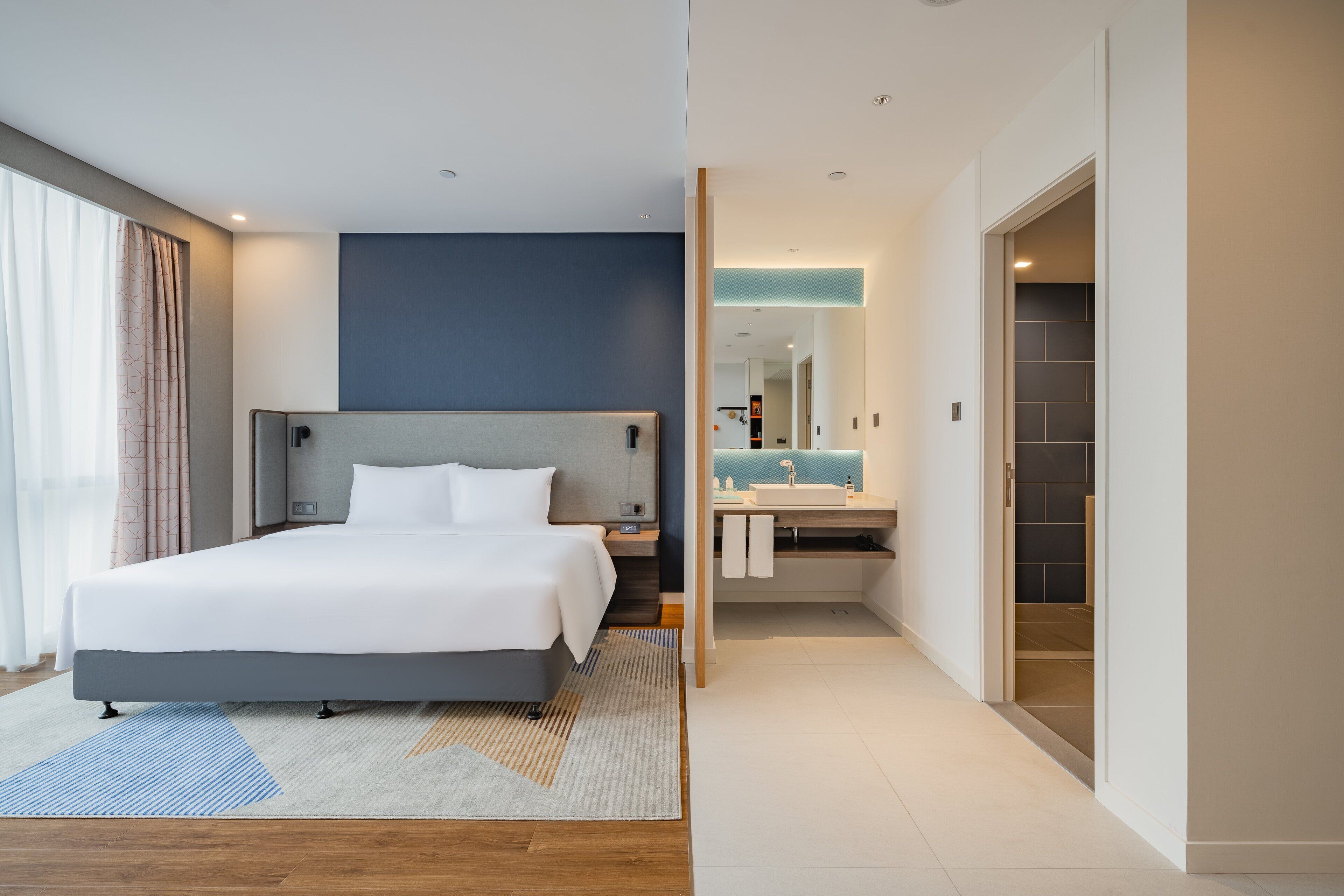 A modern hotel room with a large bed, bedside tables, a nightstand, a bathroom with a sink and mirror, and a walk-in closet.