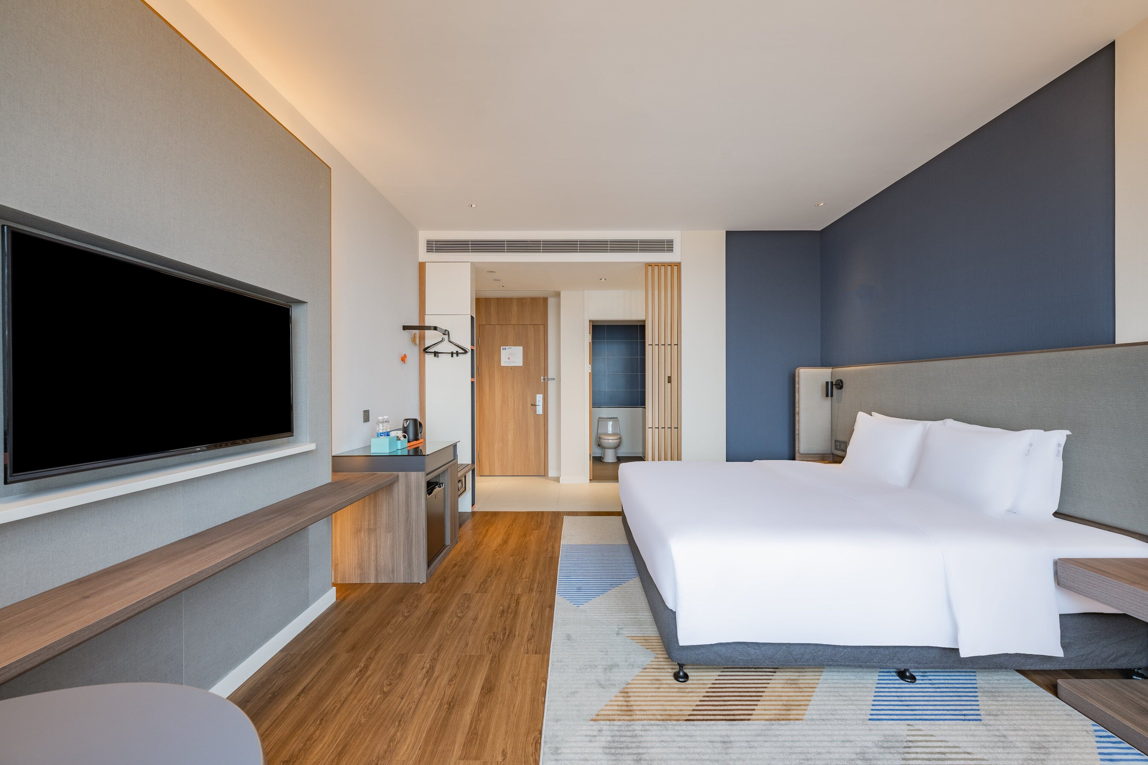 A modern hotel room with a large bed, a flat-screen TV mounted on the wall, a desk with a chair, and a bathroom visible through an open door.