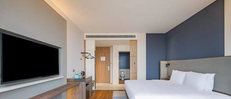 A modern hotel room with a large bed, a flat-screen TV mounted on the wall, a desk with a chair, and a bathroom visible through an open door.
