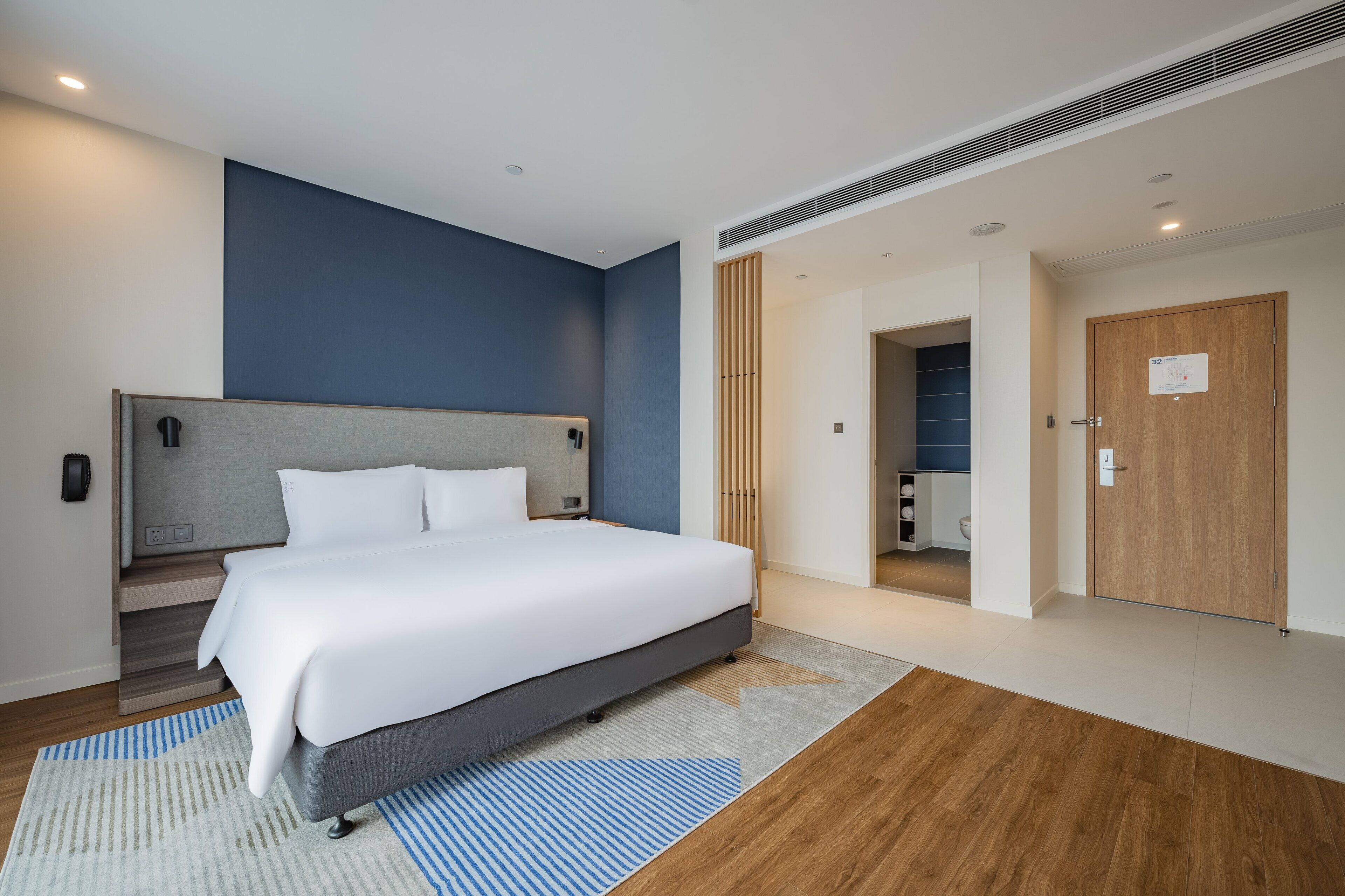 Une chambre d’hôtel moderne avec un grand lit, une table de chevet, une armoire intégrée et une salle de bain visible à travers une porte ouverte.