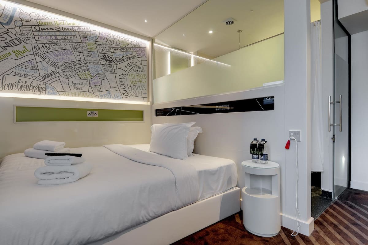 A modern hotel room with a neatly made bed, a map on the wall, a small bedside table with bottles, and a shower area.