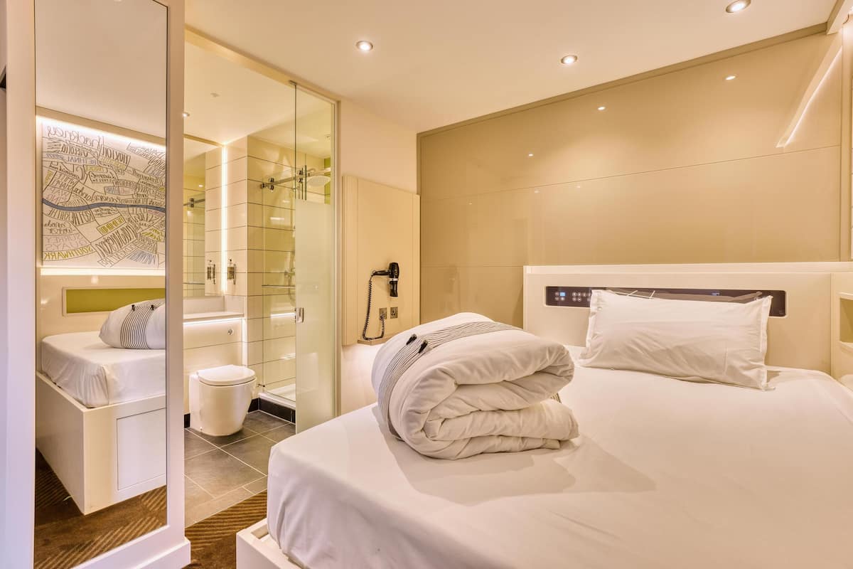 A modern hotel room with a bed, a bedside table, a mirror, a glass shower enclosure, and a map on the wall.