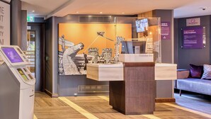 A hotel reception area with a digital kiosk, a reception desk, a sofa, and a wall-mounted TV. - Premier Inn Oxford City Centre - Westgate (Oxford)