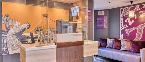 A hotel reception desk with a digital screen, a glass partition, and a sofa with patterned cushions.
