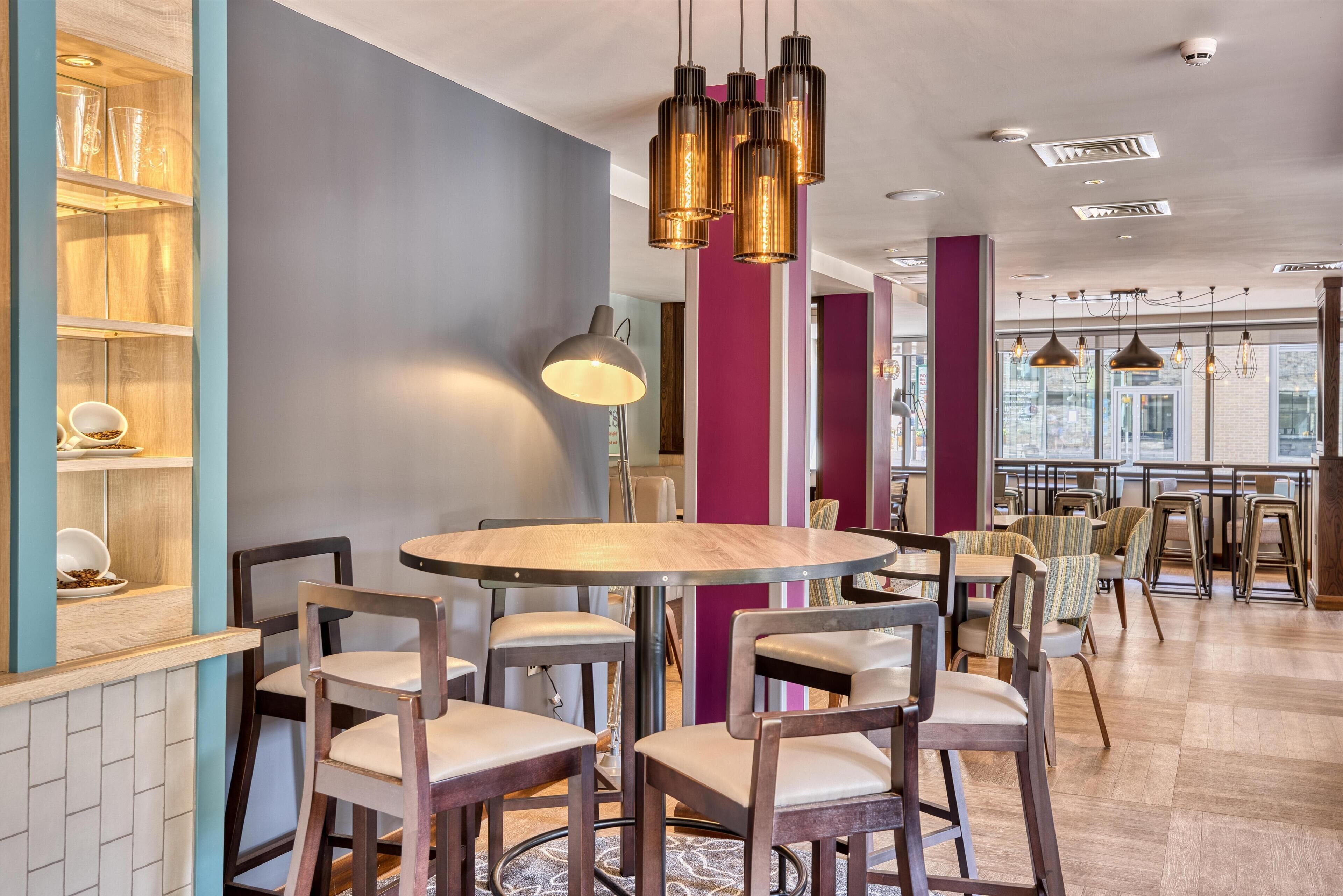 Un área de comedor moderna con mesas redondas, sillas, iluminación colgante y vistas de la ciudad a través de amplios ventanales.