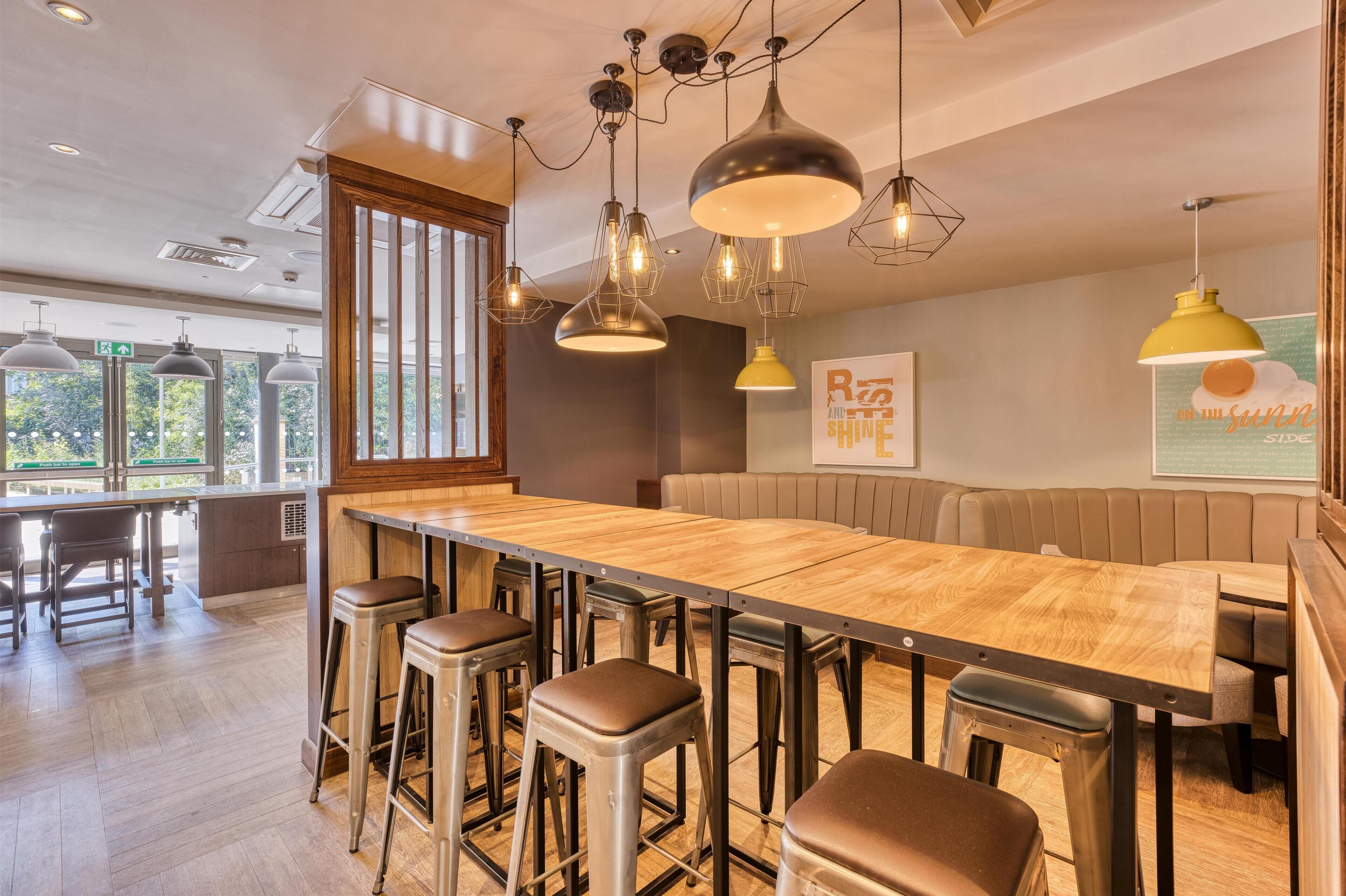 Una zona de bar moderna con una larga barra de madera, taburetes metálicos e iluminación colgante.