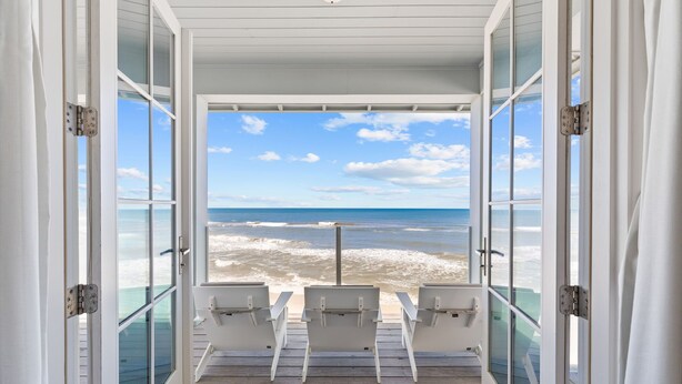 Une vue sur la plage depuis une terrasse couverte avec des portes vitrées, deux chaises blanches et une rambarde.