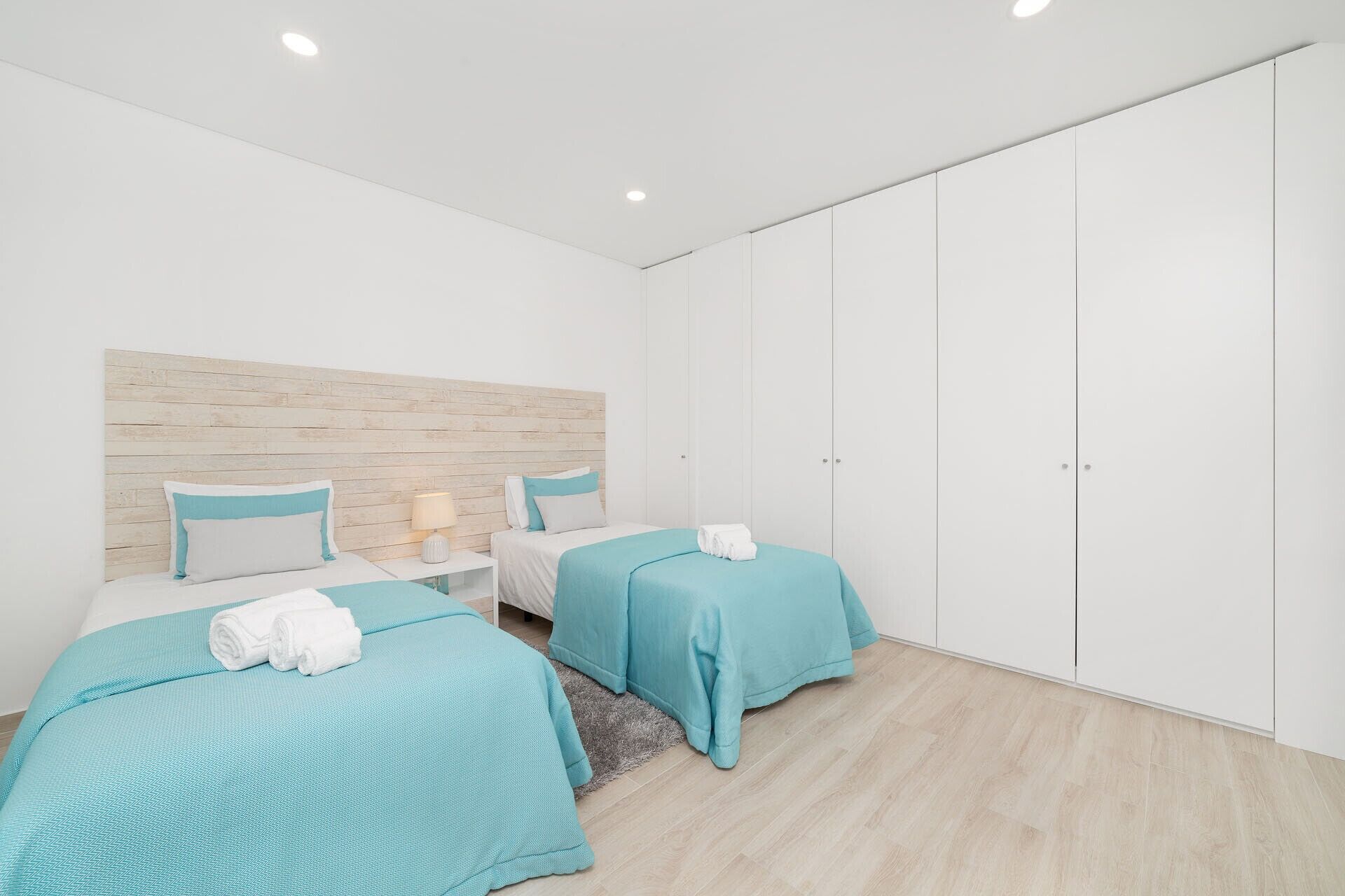 A modern bedroom with two beds, a wooden headboard, a white wardrobe, and a small nightstand with a lamp.