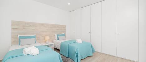 A modern bedroom with two beds, a wooden headboard, a white wardrobe, and a small nightstand with a lamp.