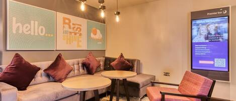 A hotel lobby with a sofa, two round tables, a chair, and a television screen displaying a message.
