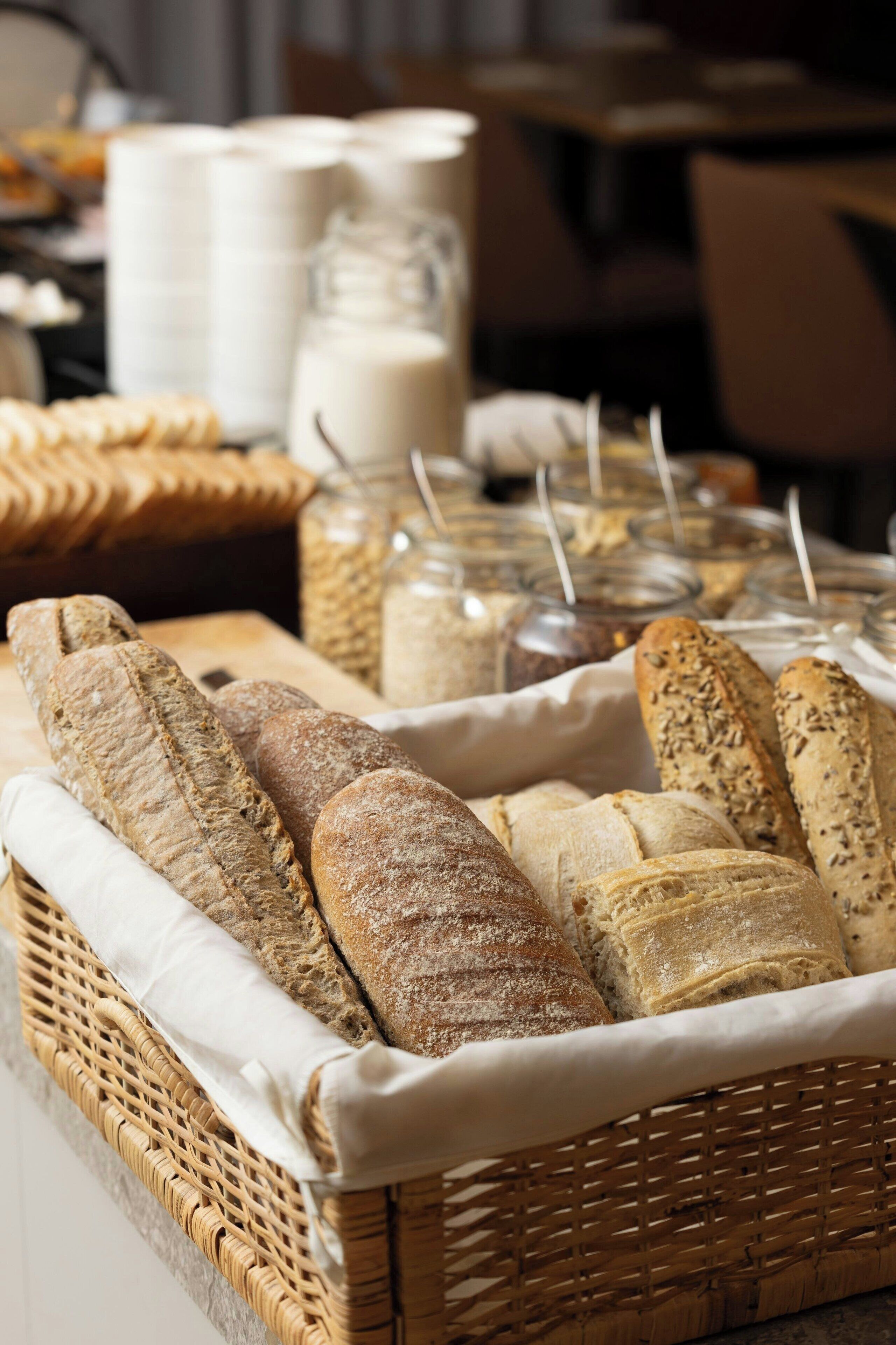 A variety of breads in a wicker basket, with jars of spreads and milk in the background.