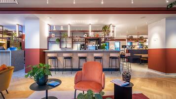 Un vestíbulo de hotel moderno con una zona de bar central, diferentes espacios de asiento y una combinación de suelos de madera y alfombra.