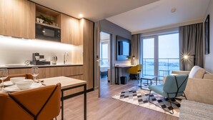 Habitación de hotel moderna con zona de comedor, sofá, televisión y balcón con vistas.