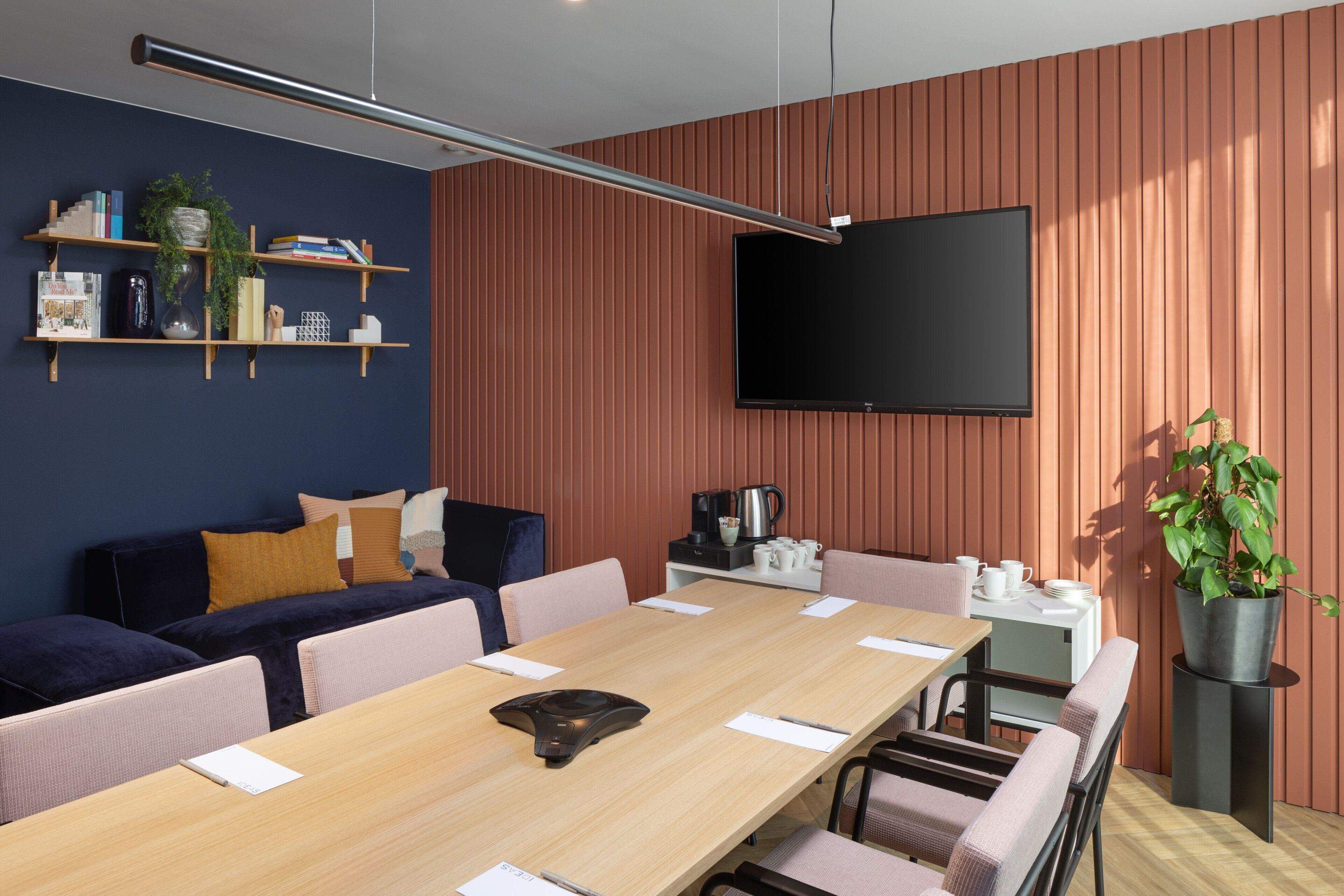 Una sala de reuniones moderna con una mesa larga, sillas, un sofá, un televisor, estanterías con libros y plantas, y un rincón para café.