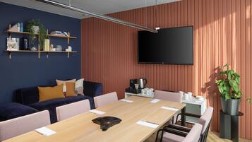 Una sala de reuniones moderna con una mesa larga, sillas, un sofá, un televisor, estanterías con libros y plantas, y un rincón para café.