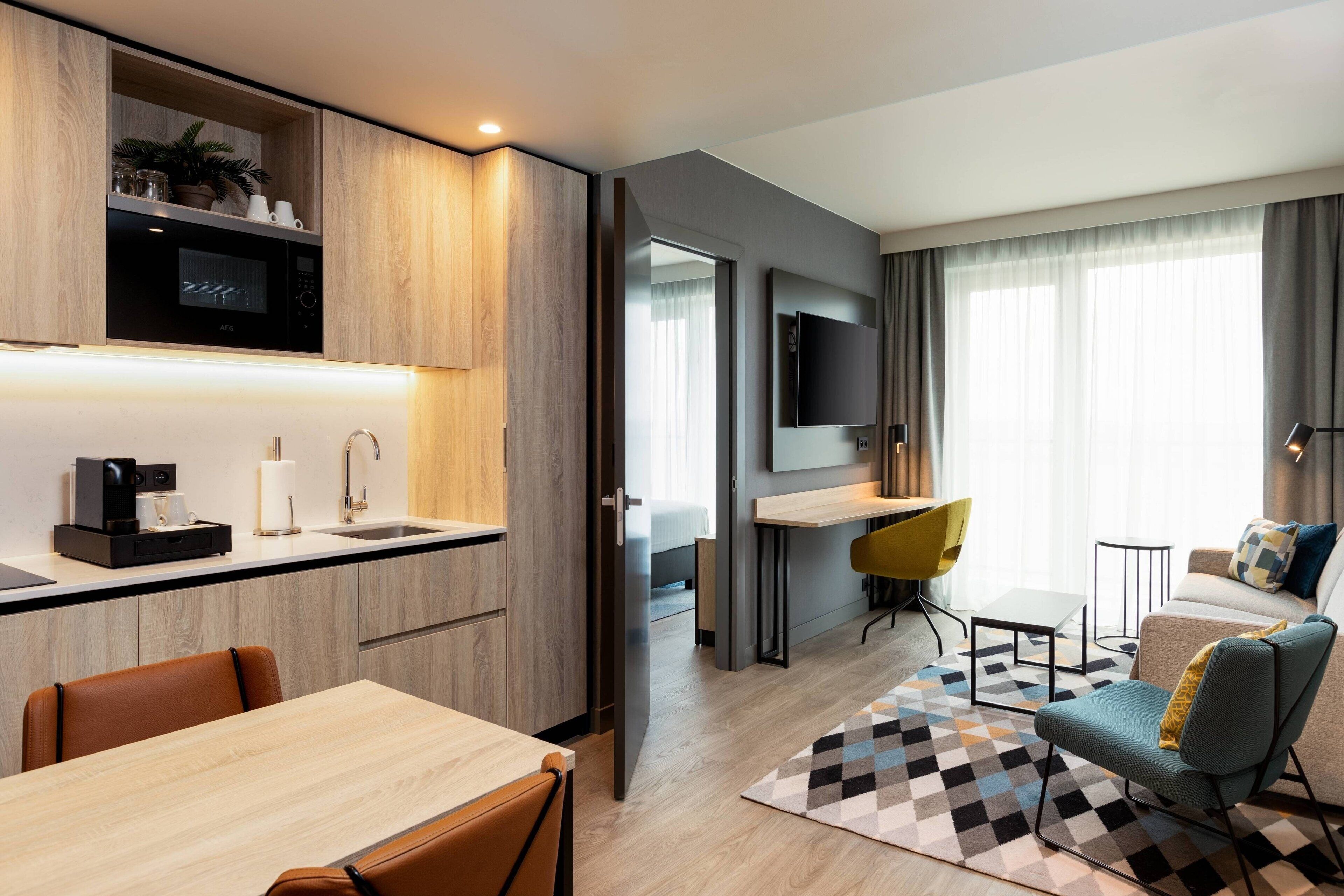A modern hotel room with a kitchenette, a dining area, a living space with a sofa and armchair, and a large window with curtains.
