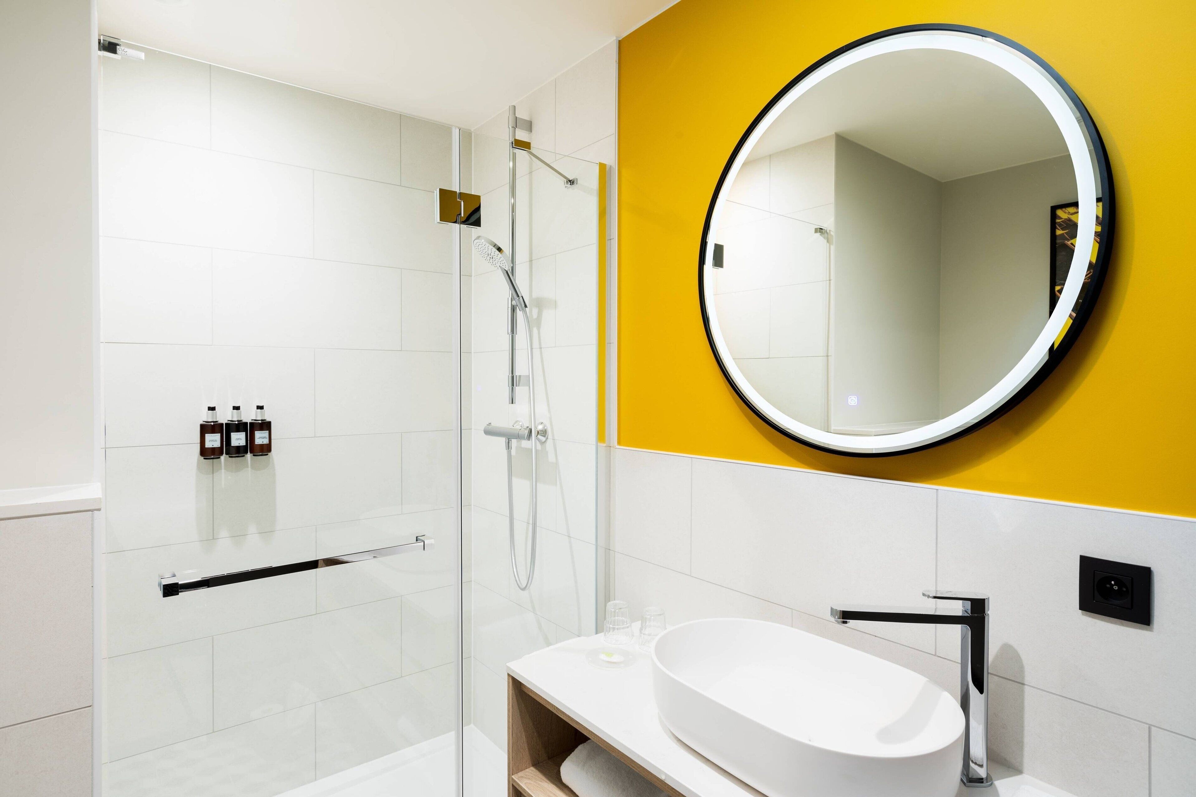 A modern bathroom with a glass-enclosed shower, a white sink with a chrome faucet, and a round mirror with a black frame.