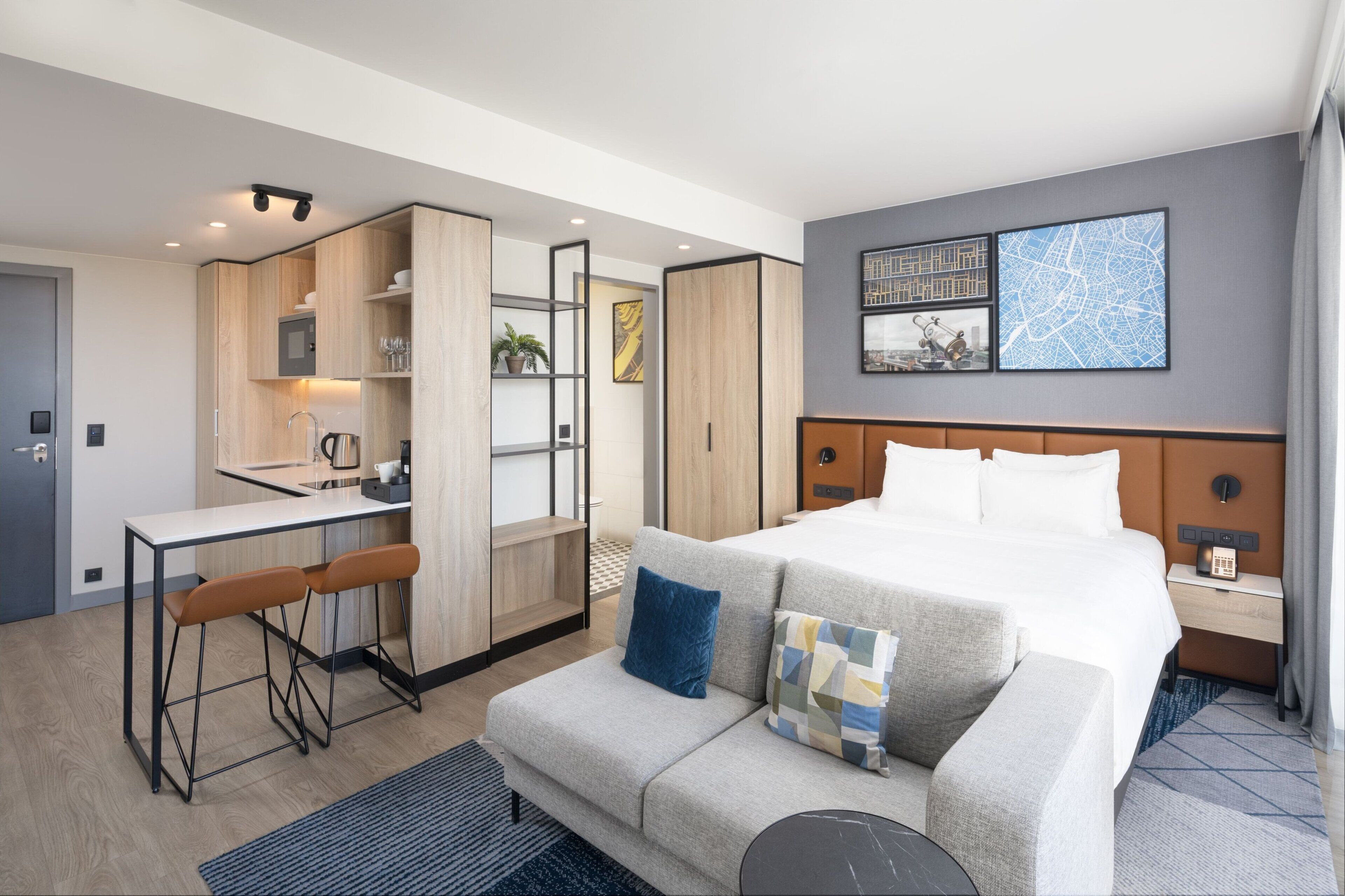 A modern hotel room with a bed, a sofa, a desk, and a kitchenette.