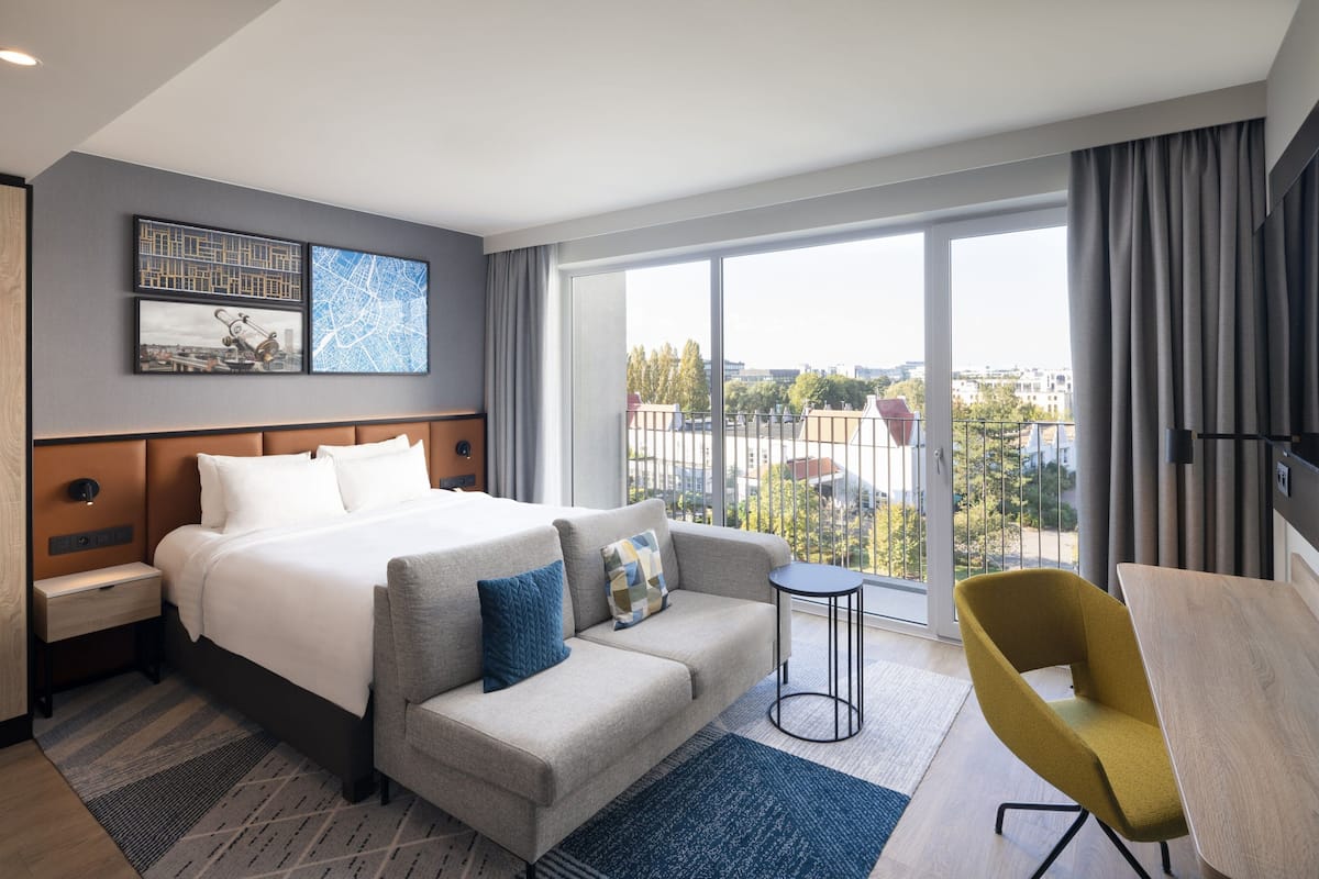 Ein Hotelzimmer mit Bett, Sofa, kleinem Tisch und Stuhl. Zwei Fenster mit Vorhängen bieten Blick auf die Stadt.