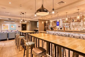 Una zona de bar con mesas y taburetes de madera, una barra con botellas y vasos, y una pared con un letrero.