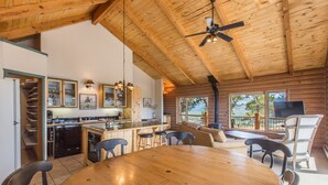 A spacious cabin interior with a wooden ceiling, a central island with stools, a dining table, and a cozy seating area with a fireplace. - Wrap Around Deck | Unmatched Views | Air Conditioning | STR2025-060 (Ridgway)