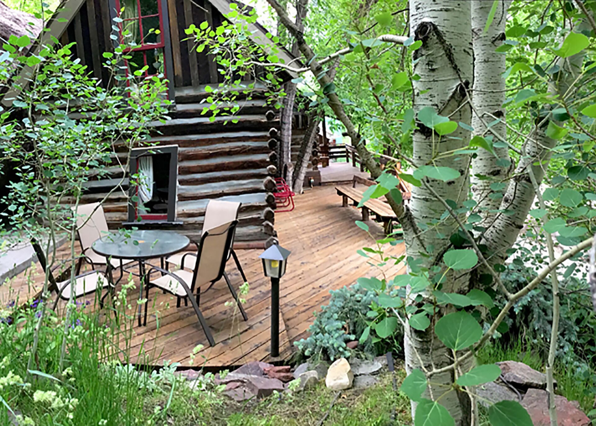 A wooden deck with a table and chairs, surrounded by greenery and a log cabin in the background.