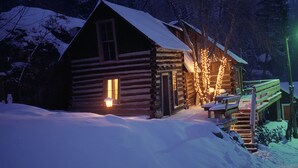 Een blokhut met een met sneeuw bedekte dak, een houten trap en een veranda met lichtjes.