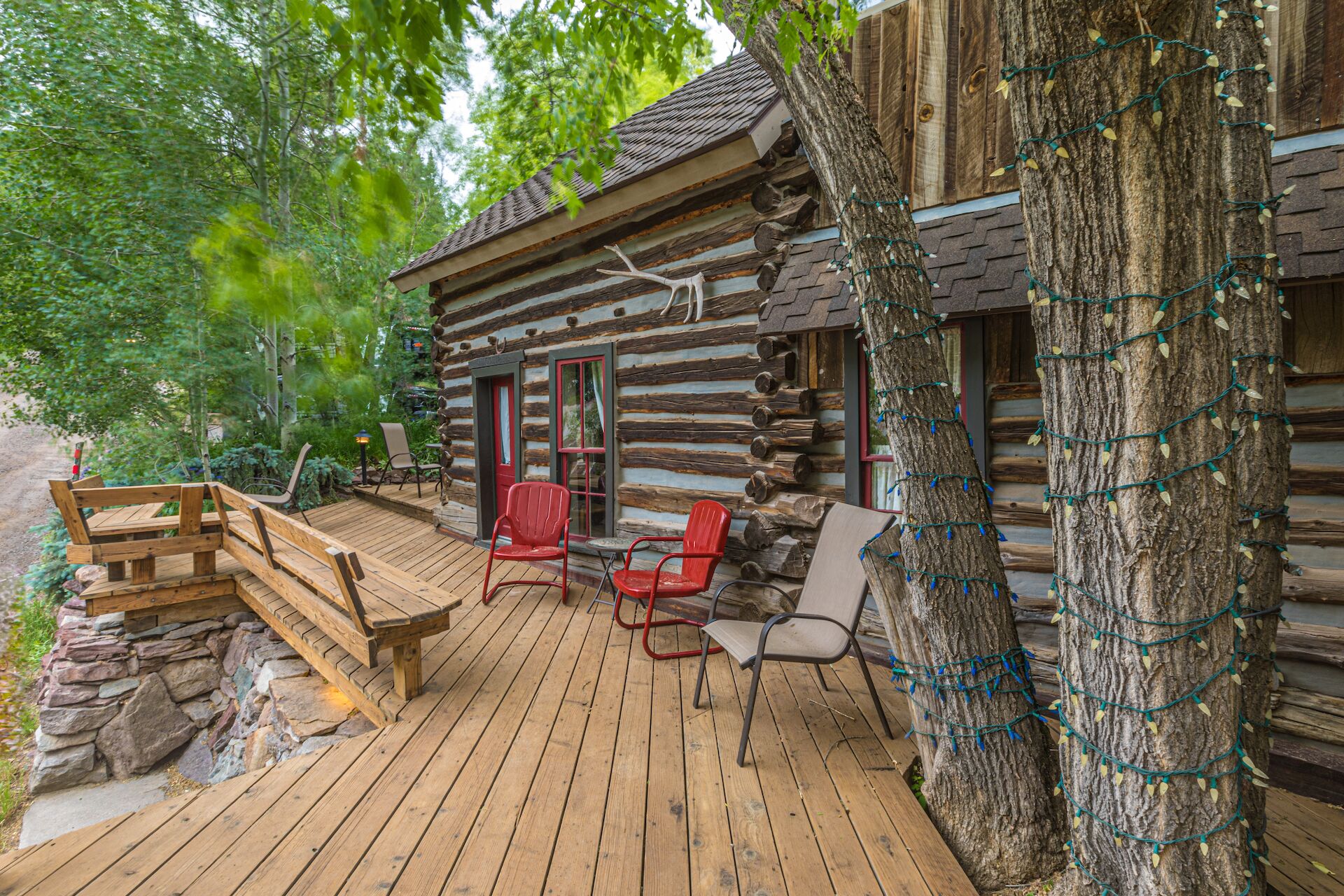 Une cabane rustique en rondins de bois, dotée d’une terrasse en bois, de deux chaises rouges et d’un banc.