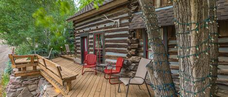 Une cabane rustique en rondins de bois, dotée d’une terrasse en bois, de deux chaises rouges et d’un banc.