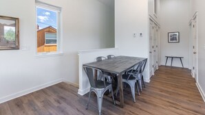 Een eetgedeelte met een houten tafel en metalen stoelen, witte muren en een raam met uitzicht op een gebouw van hout.