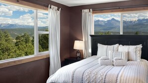 A bedroom with a bed, curtains, a nightstand, a lamp, and a view of mountains.