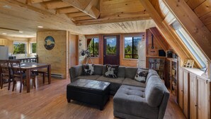 A cozy cabin interior with a large wooden ceiling, a sectional sofa, a dining area, and a view of mountains through the windows.