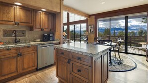 A kitchen with wooden cabinets, granite countertops, and a dishwasher. There is a large window offering a view of mountains. - Mountain Retreat | Stunning Views | A/C | Hot Tub STR2025-062 (Ridgway)