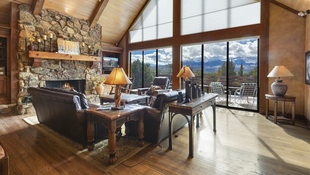 A spacious living room with a stone fireplace, wooden beams, and a large window offering a mountain view. - Mountain Retreat | Stunning Views | A/C | Hot Tub STR2025-062 (Ridgway)