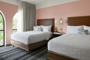 Quarto de hotel com duas camas, uma janela grande com cortinas, um tapete estampado e uma mesa de cabeceira com abajur.