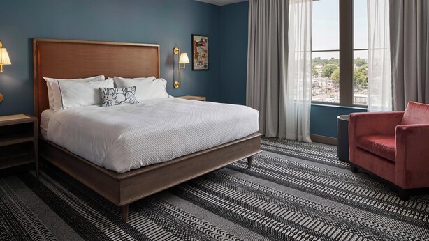 A hotel room with a large bed, a red armchair, a nightstand with a lamp, and a view of the city through the window.