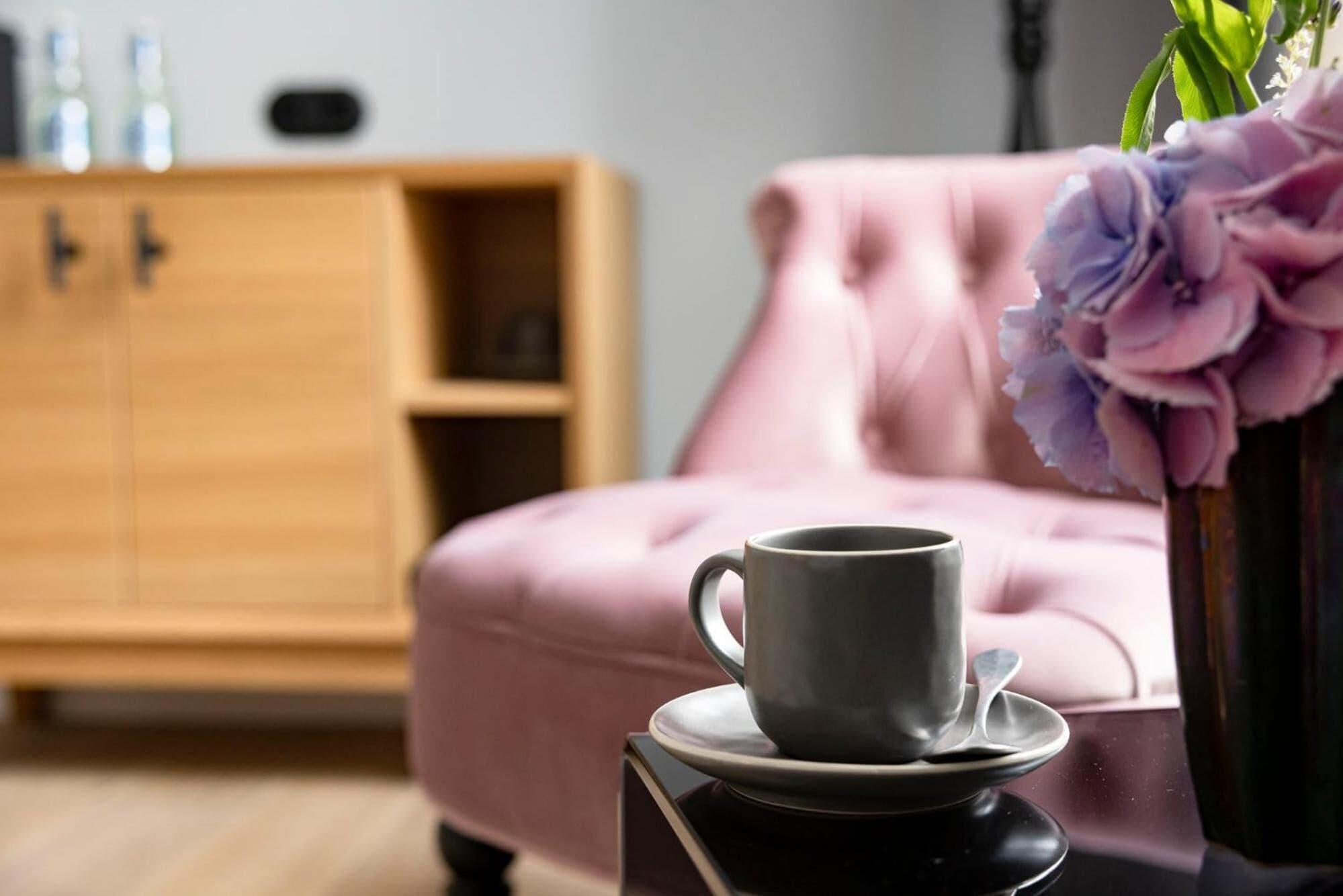 Um quarto acolhedor com um sofá rosa, uma mesinha com xícara e pires, e um armário de madeira ao fundo.