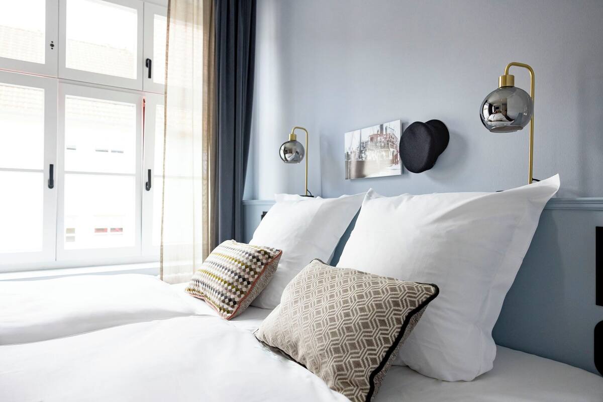 Een slaapkamer met een bed, kussens, een wandlamp en een raam met gordijnen.